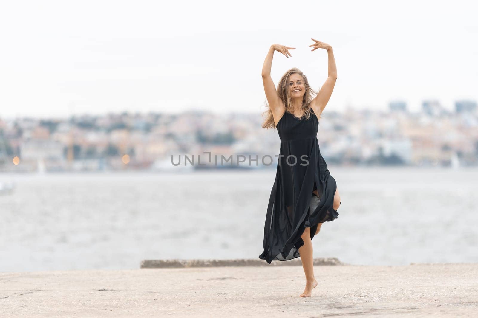 An adult smiling woman in black dress dancing ballet on the pier in cloudy weather. Mid shot