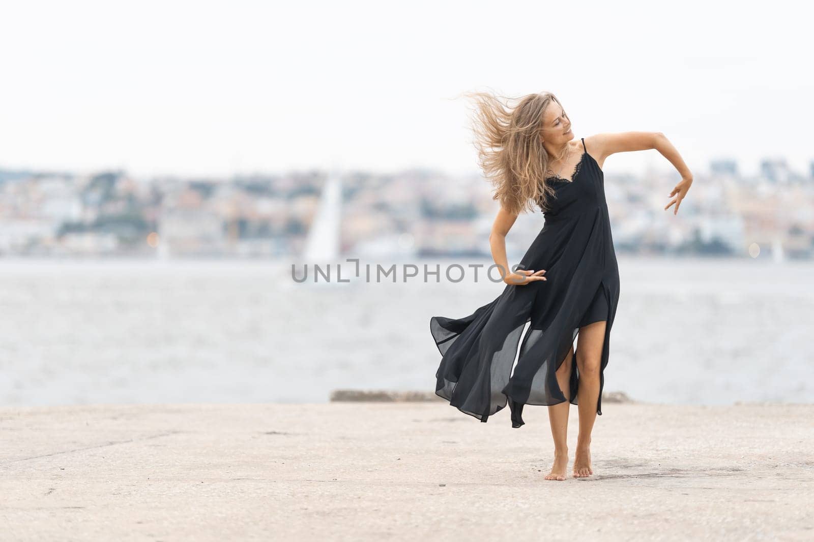 An adult smiling woman in black dress dancing ballet on the pier. Mid shot