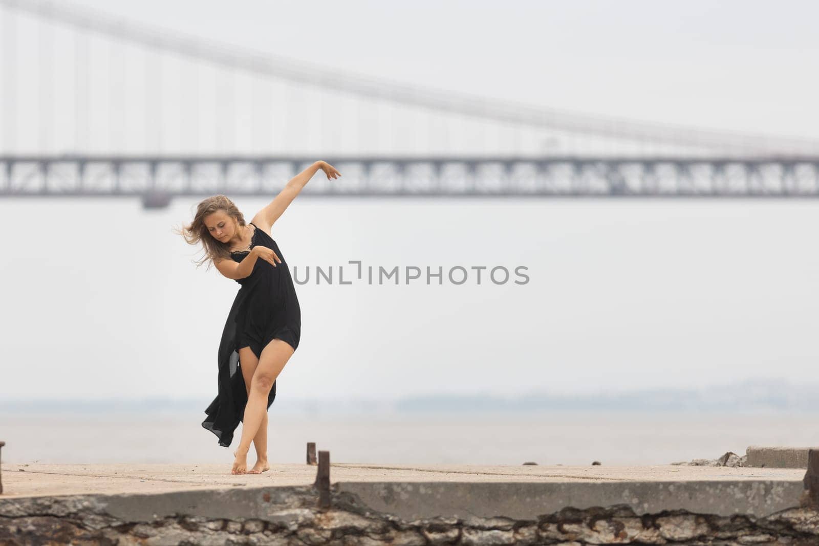 An adult woman in black dress dancing ballet on the pier with a bridge on the background. Mid shot
