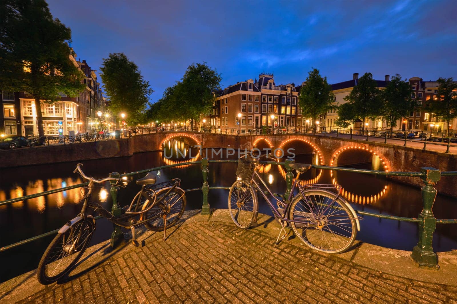 Night view of Amsterdam cityscape with canal, bridge with bicycles and medieval houses in the evening twilight illuminated. Amsterdam, Netherlands