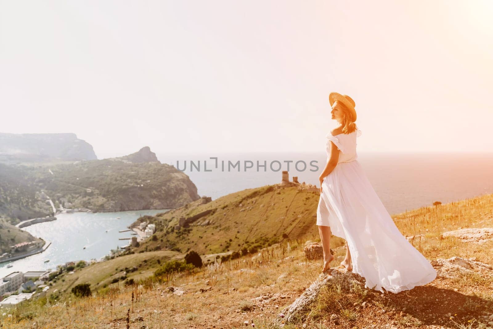 Happy woman in a white dress and hat stands on a rocky cliff above the sea, with the beautiful silhouette of hills in thick fog in the background. by Matiunina