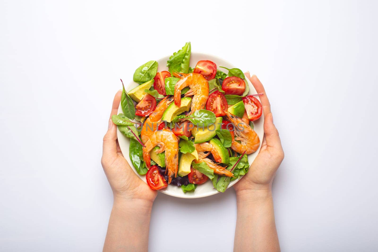 Female hands holding healthy salad with grilled shrimps, avocado, cherry tomatoes and green leaves on white plate isolated on white background top view. Clean eating, nutrition and dieting concept..