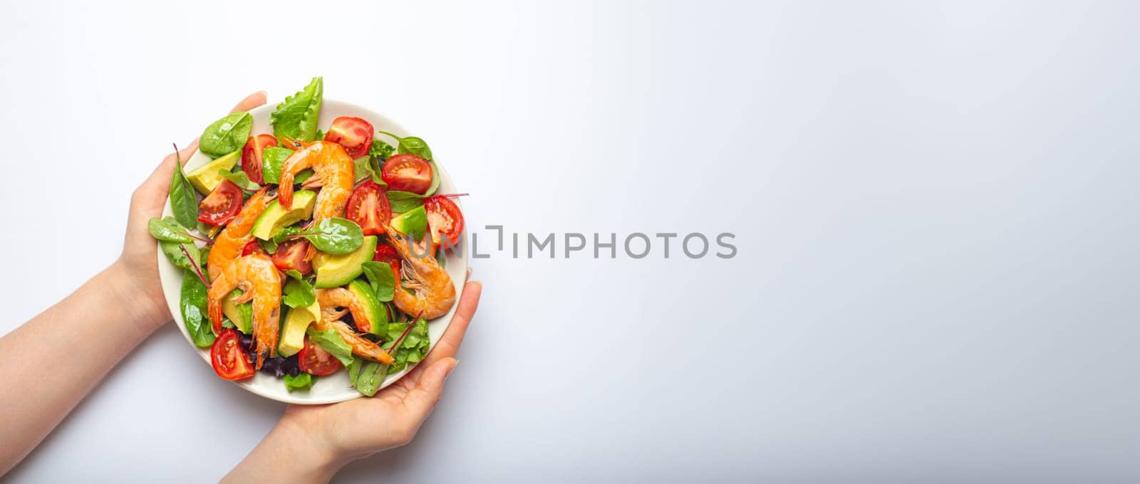 Female hands holding salad with grilled shrimps, avocado, vegetables, green leaves on white plate isolated on white background top view. Clean eating, nutrition and dieting concept, copy space..