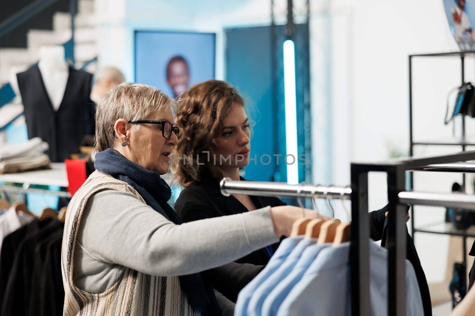 Showroom employee helping elderly woman choosing trendy clothes, discussing fabric in modern boutique. Senior client shopping for casual wear, buying fashionable merchandise in clothing store