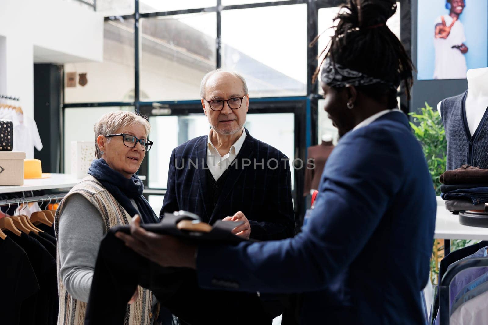 Elderly couple discussing shirt fabric with showroom worker, buying trendy clothes for formal wear in modern boutique. Senior man looking at new fashion collection in shopping centre. Fashion concept