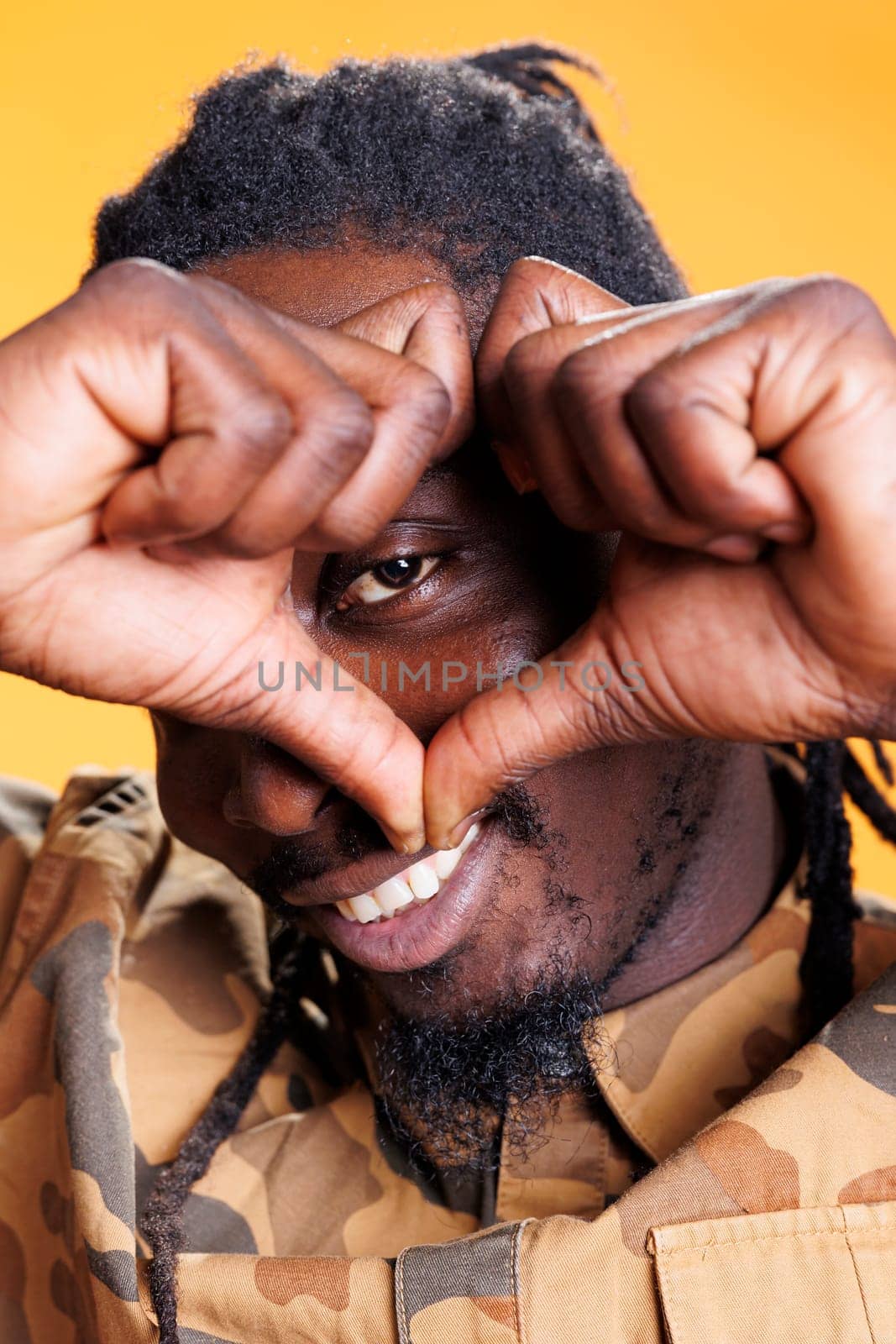 Lovely person doing heart shape in front of camera, smiling during valentine s day studio shot. African american man making love symbol with fingers over yellow background. Romantic gesture