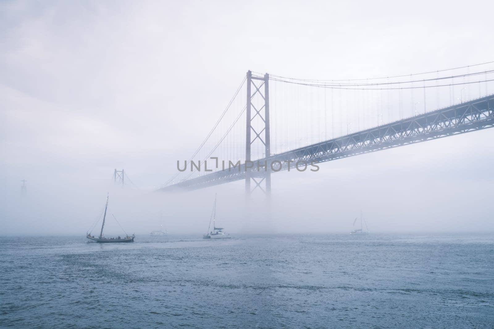 View of 25 de Abril Bridge famous tourist landmark of Lisbon connecting Lisboa and Almada in heavy fog mist wtih yacht boats passing under. Lisbon, Portugal