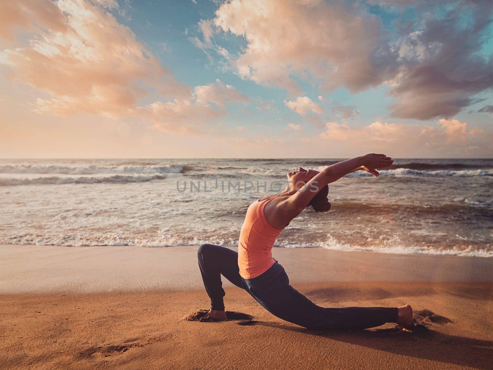 Sporty fit woman practices yoga Anjaneyasana at beach on sunset by dimol