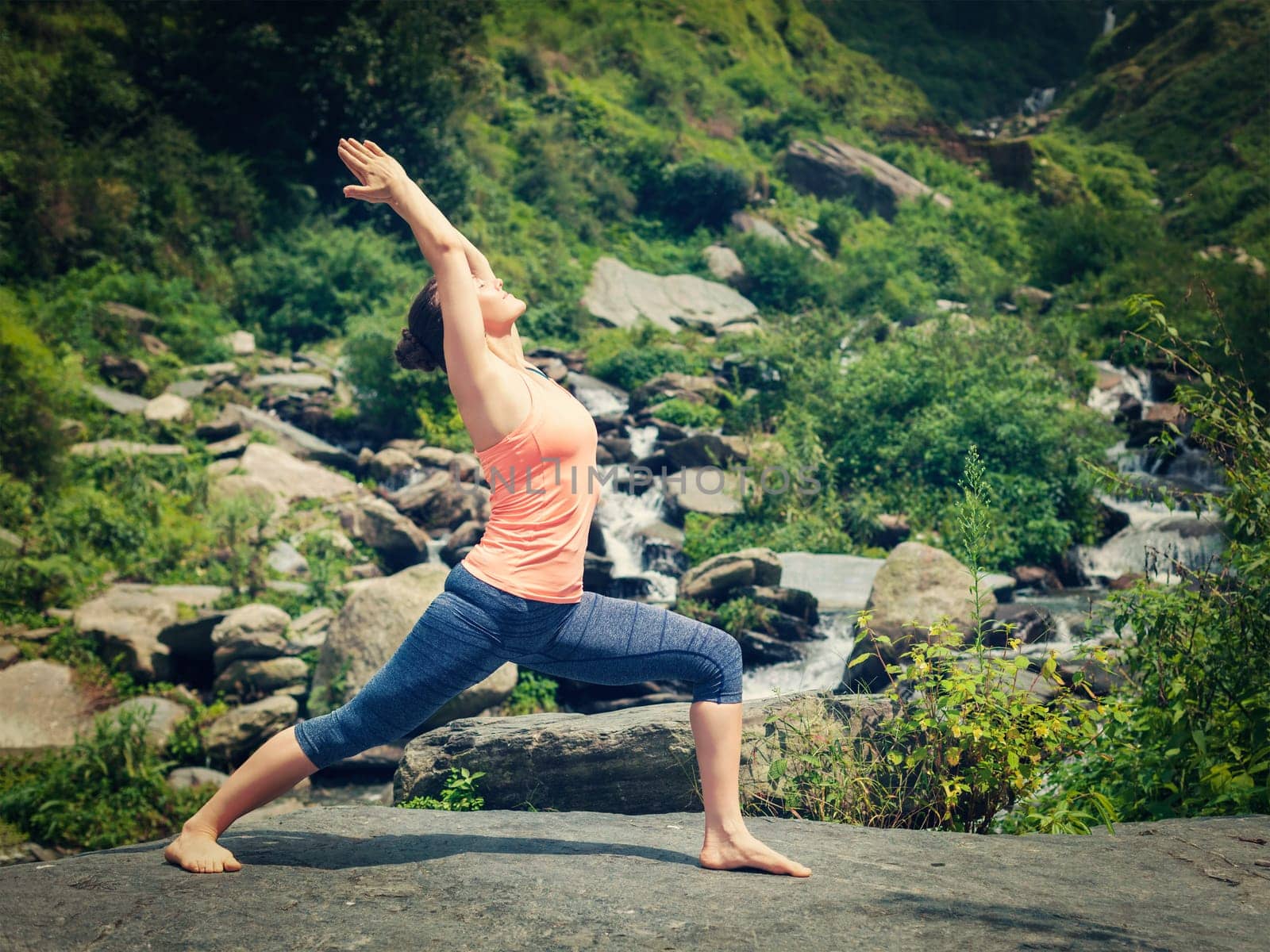 Yoga outdoors - sporty fit woman doing Ashtanga Vinyasa Yoga asana Virabhadrasana 1 Warrior pose posture at waterfall in HImalayas mountains. Vintage retro effect filtered hipster style image.