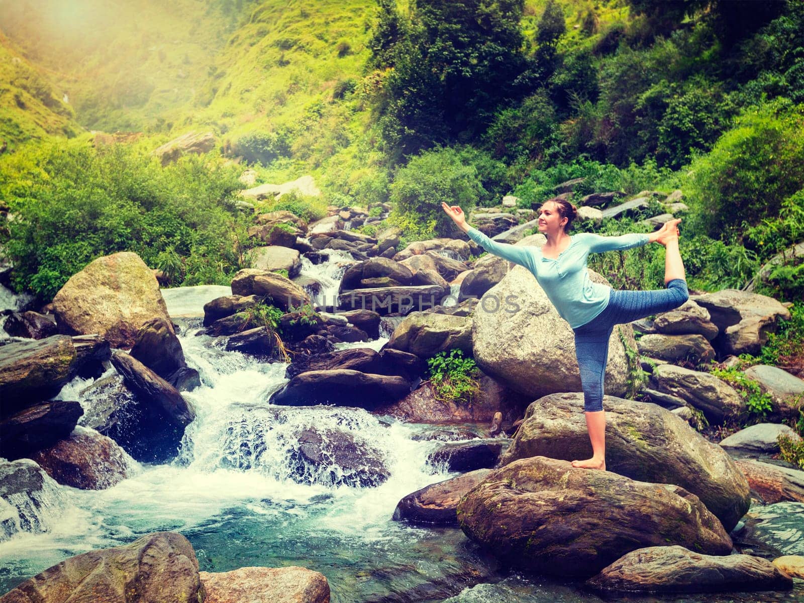 Yoga outdoors - woman doing yoga asana Natarajasana - Lord of the dance balance pose outdoors at waterfall in Himalayas. Vintage retro effect filtered hipster style image.