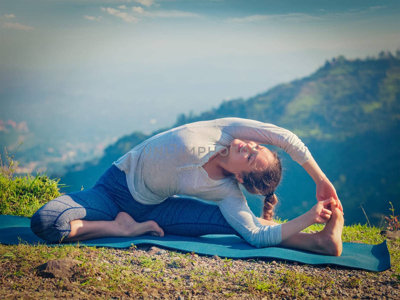 Yoga outdoors - young sporty fit woman doing Hatha Yoga asana parivritta janu sirsasana - Revolved Head-to-Knee Pose - in mountains in the morning. Vintage retro effect filtered hipster style image.