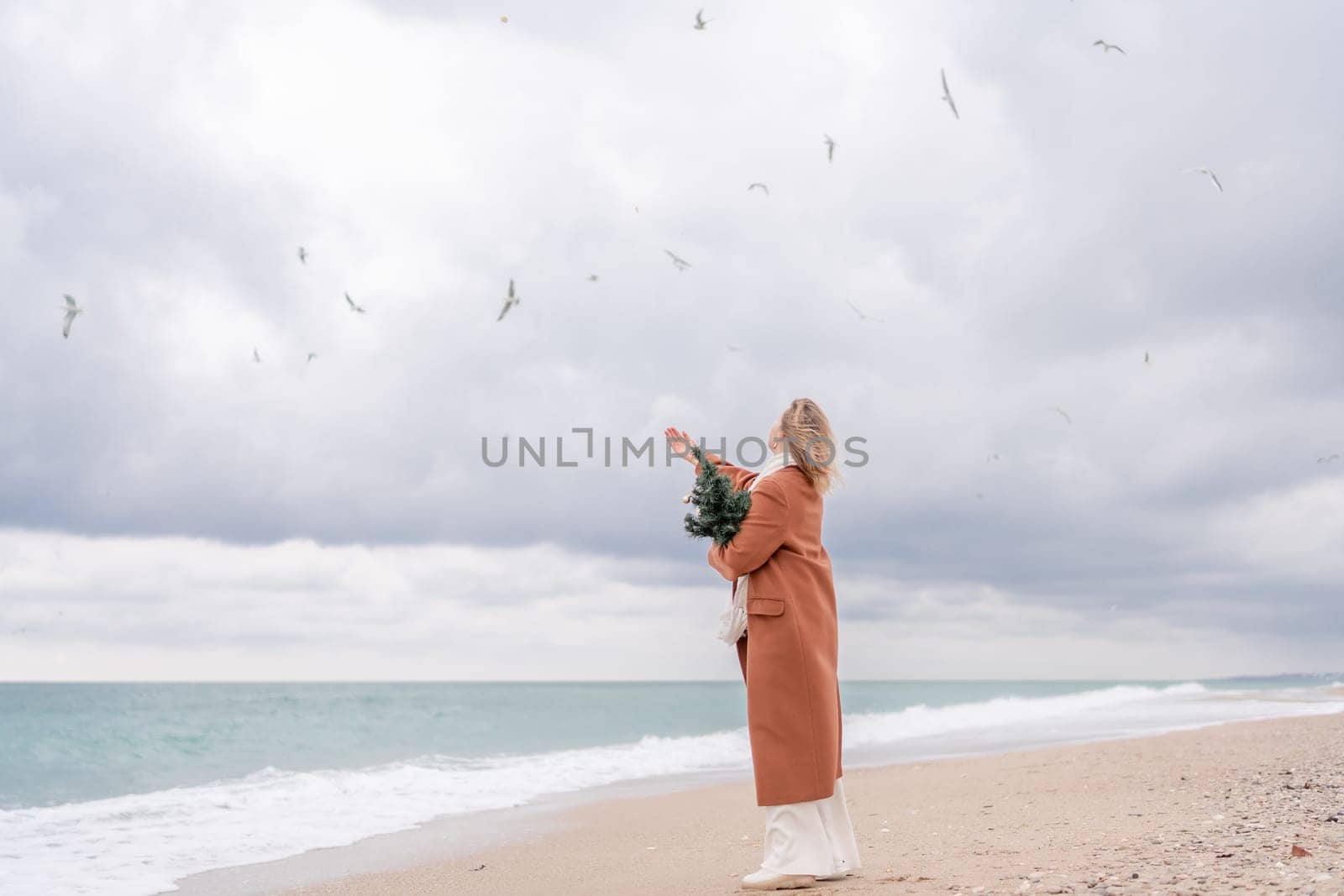 Blond woman Christmas sea. Christmas portrait of a happy woman walking along the beach and holding a Christmas tree on her shoulder. She is wearing a brown coat and a white suit