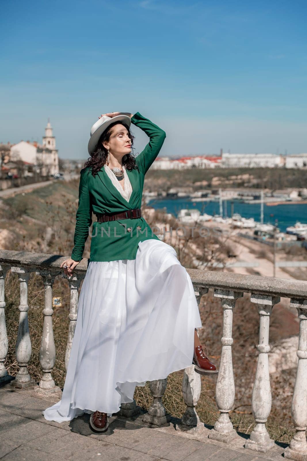 Woman walks around the city, lifestyle. A young beautiful woman in a green jacket, white skirt and hat is sitting on a white fence with balusters overlooking the sea bay and the city
