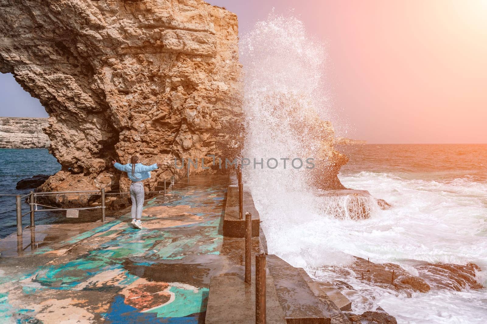 woman sea travel. A woman in a blue jacket stands on a rock above a cliff above the sea, looking at the stormy ocean. Girl traveler rests, thinks, dreams, enjoys nature.