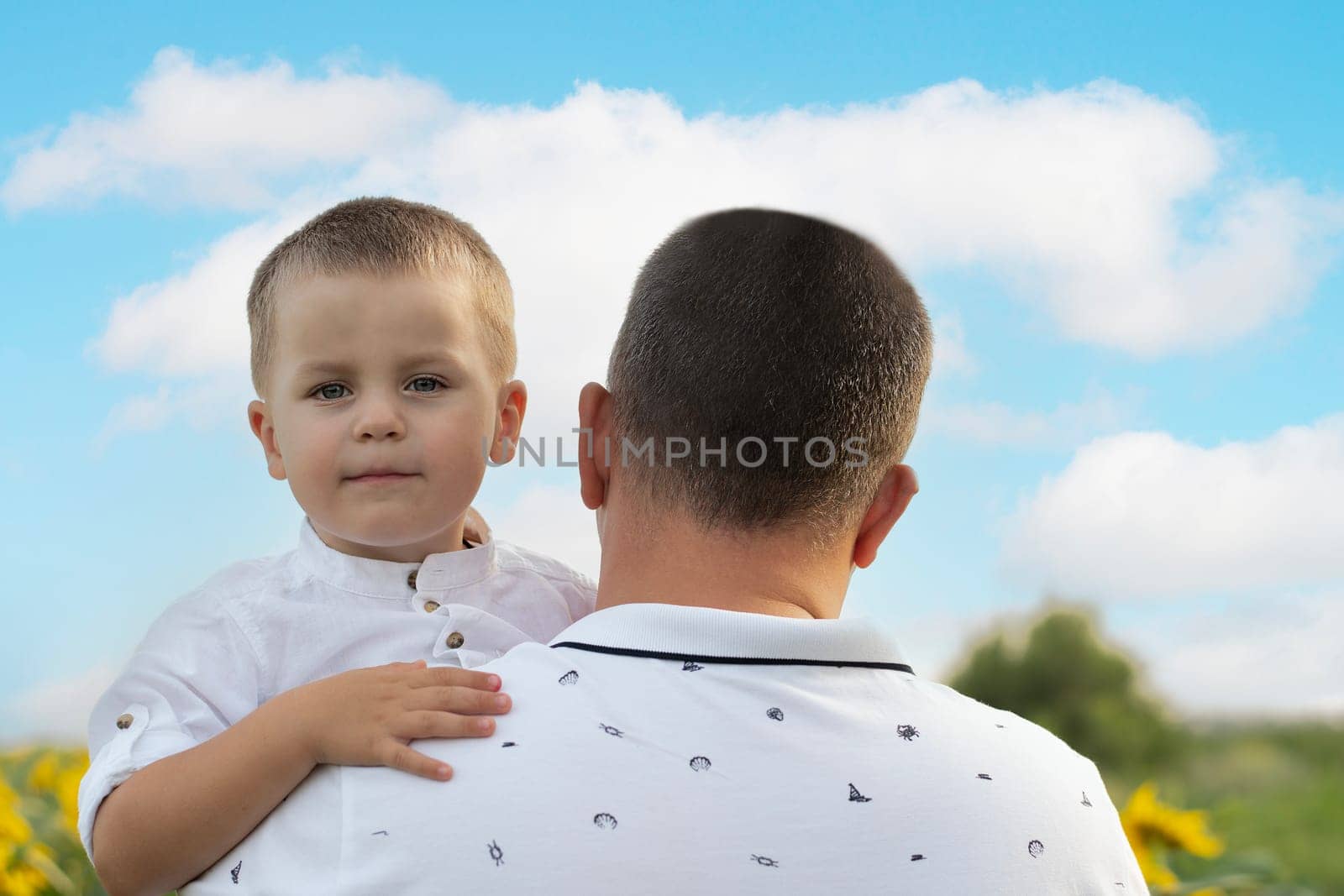 Father's day concept. A little boy in his father's arms gently hugs and cuddles, standing in a field with yellow sunflowers against the background of a blue sky. A symbol of peace. Symbol of Ukraine. by ketlit