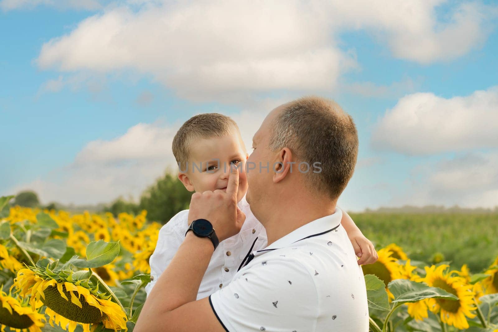 Father's day concept. A little boy in his father's arms against the blue sky stands in a field with yellow sunflowers. A symbol of peace. Symbol of Ukraine. by ketlit