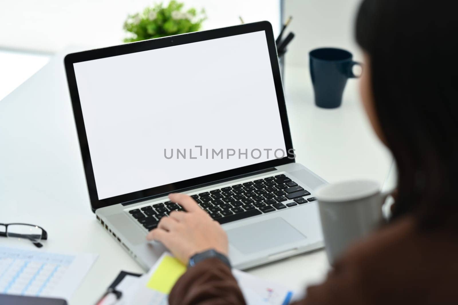 Over shoulder view of female office worker drinking coffee and using laptop at workstation.