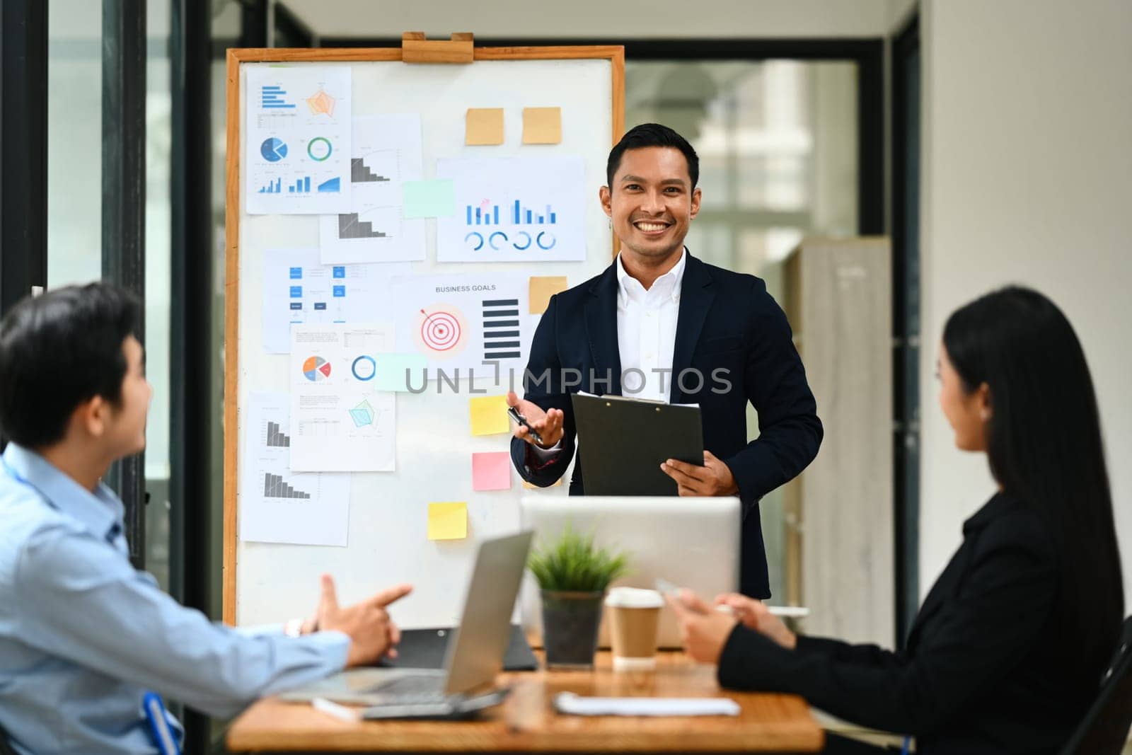 Asian male business team leader standing in front of flip chart giving presentation at meeting.