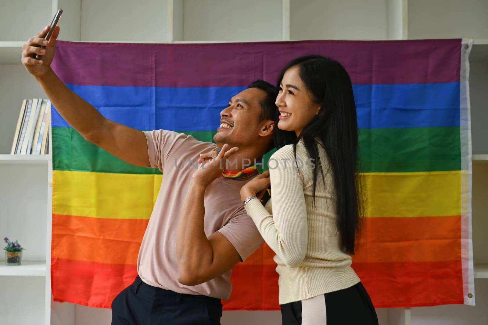 Cheerful biracial transgender man and young woman making selfie on smartphone against rainbow flag.