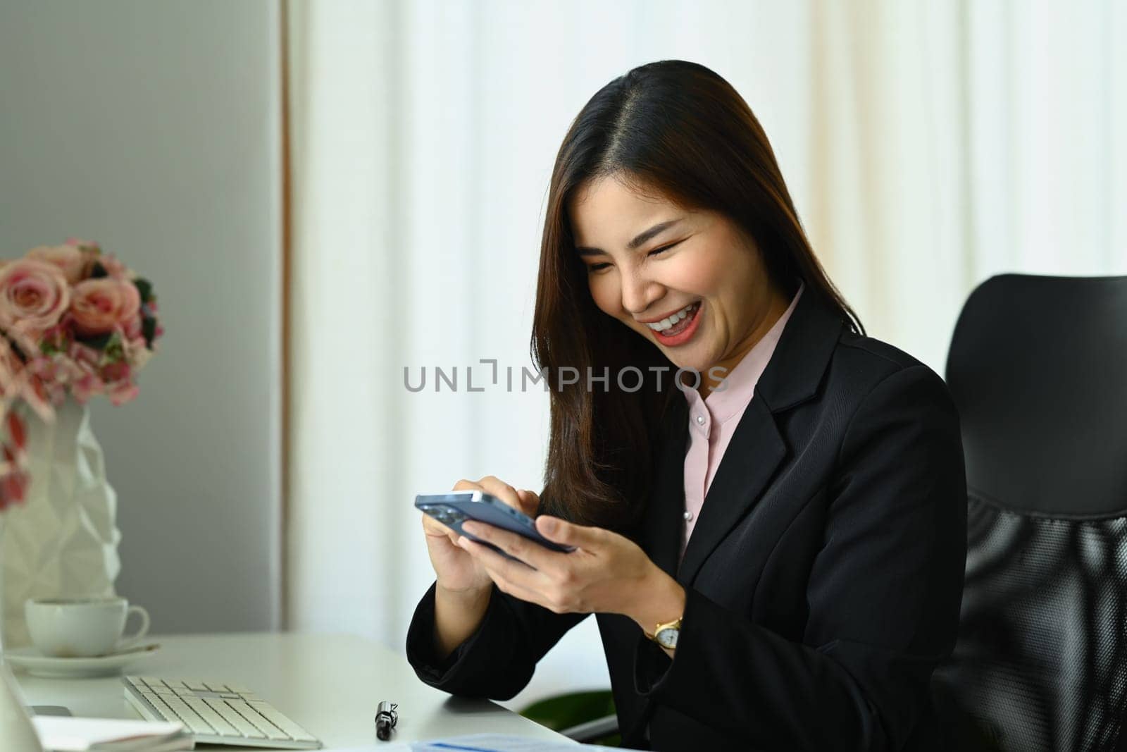 Cheerful female employee sitting at desk typing massage, communicating online on mobile phone.