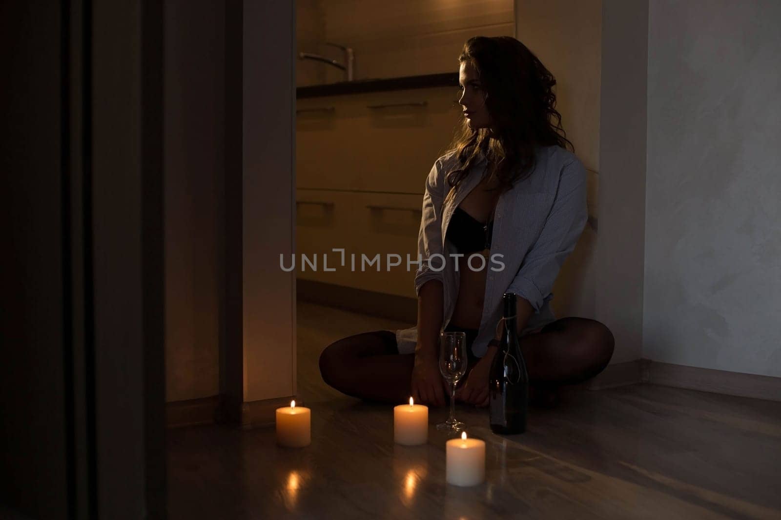 A sexy brunette in a black bra and a man's shirt sits on the floor by candlelight. seductive girl in underwear and stockings lit candles for a romantic setting by malyshph