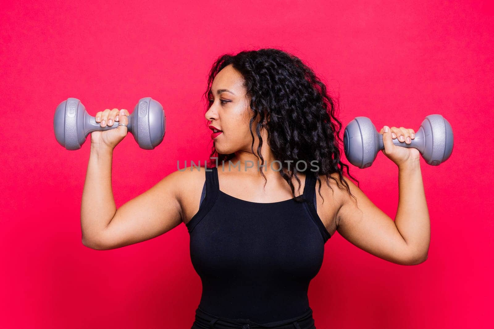 Smiling african american female doing hand exercises with dumbbells on a red studio background.