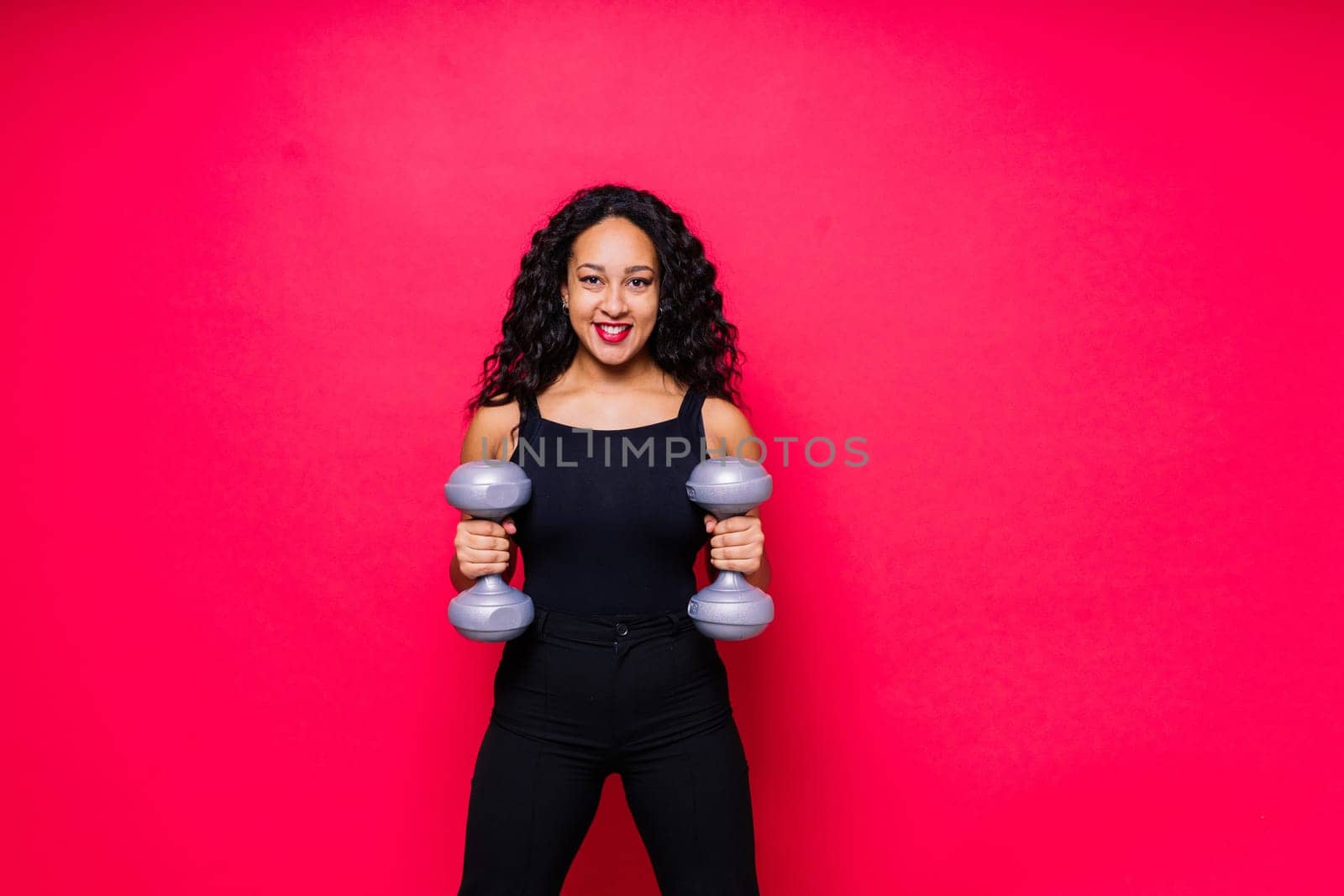 Smiling african american female doing hand exercises with dumbbells on a red studio background.