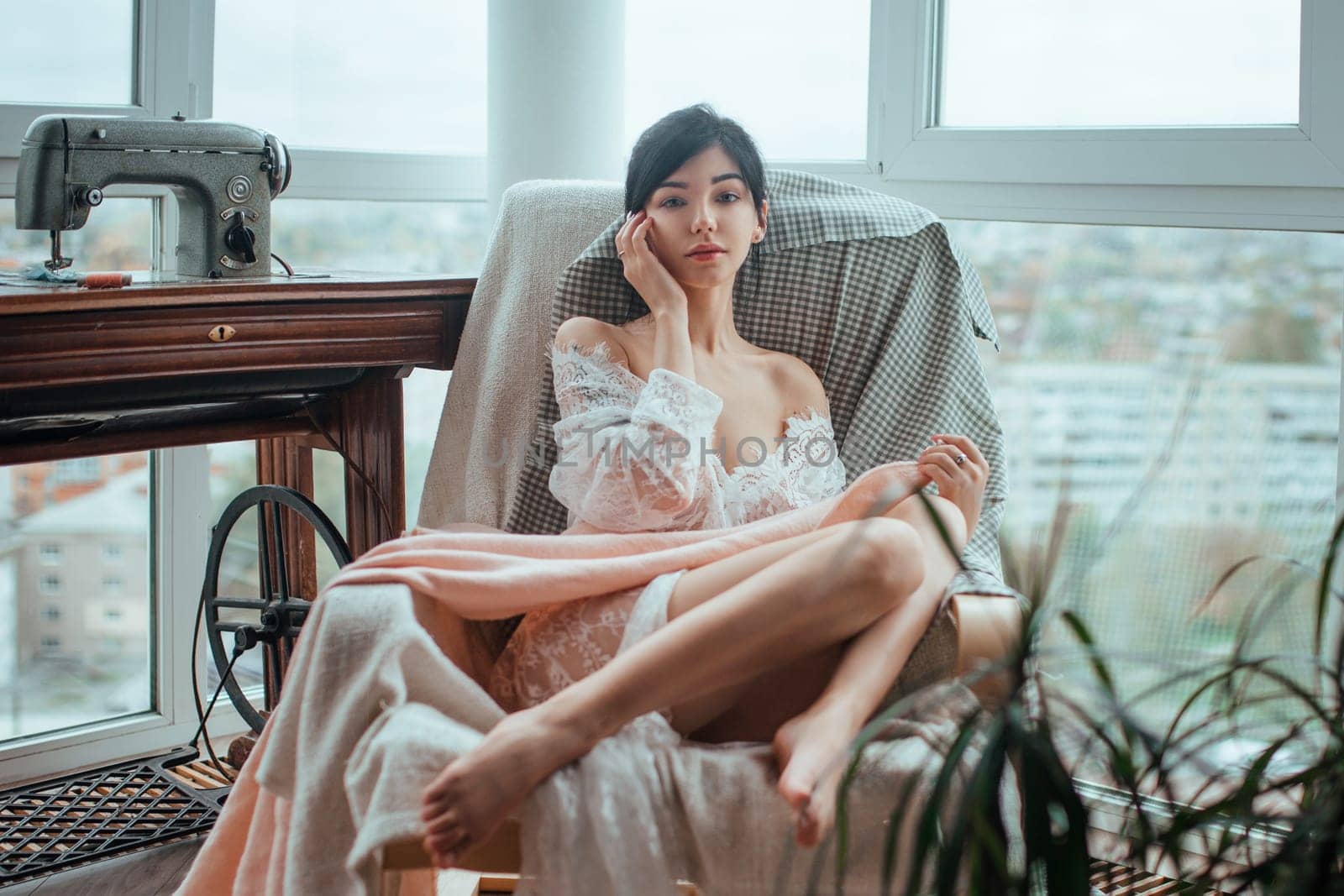 Sexy girl in a white translucent tunic sits sewing near the window.Beautiful female body shines through the fabric.