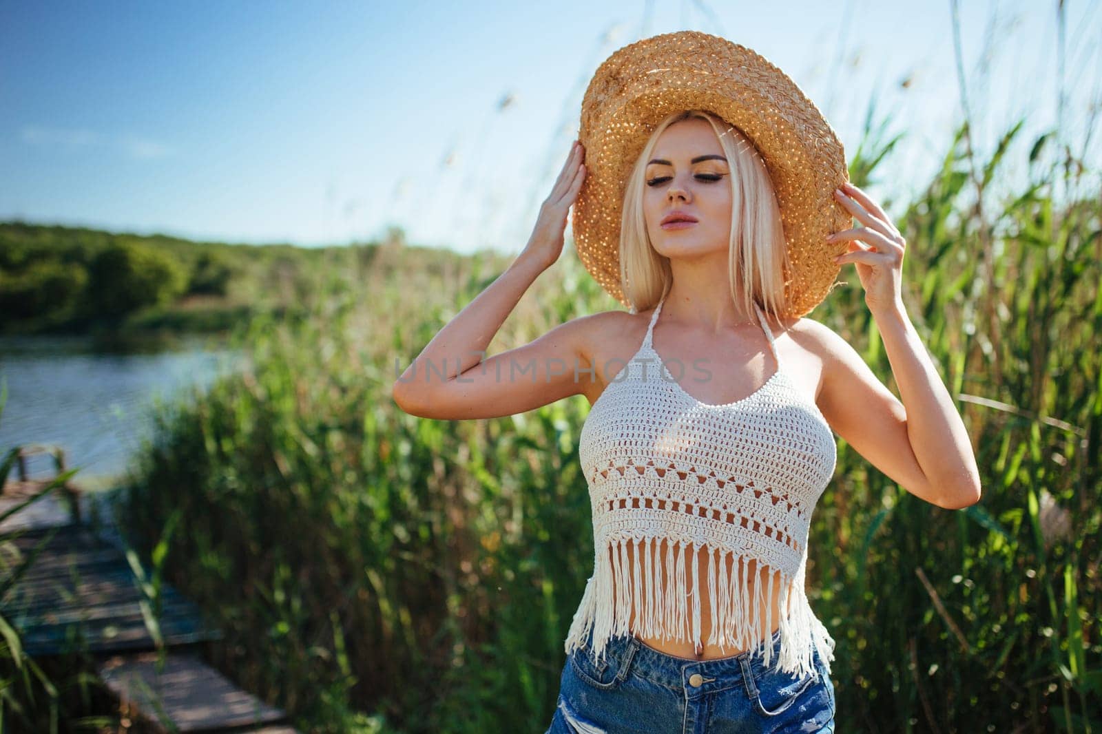 Beautiful model girl posing on the field enjoying nature outdoors in a straw hat. Beauty blond young woman with long straight blond hair close up portrait.