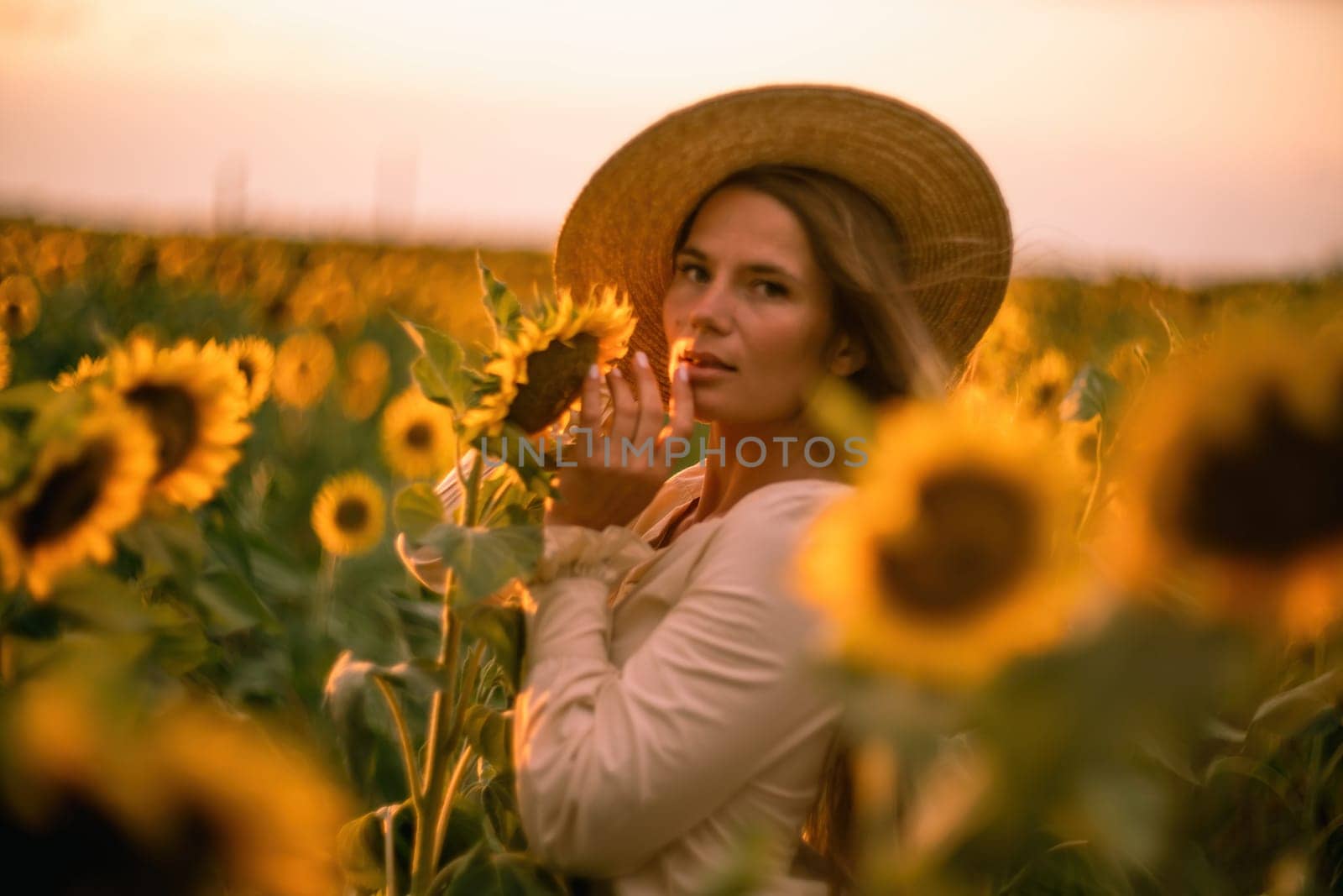 Beautiful middle aged woman looks good in a hat enjoying nature in a field of sunflowers at sunset. Summer. Attractive brunette with long healthy hair. by Matiunina