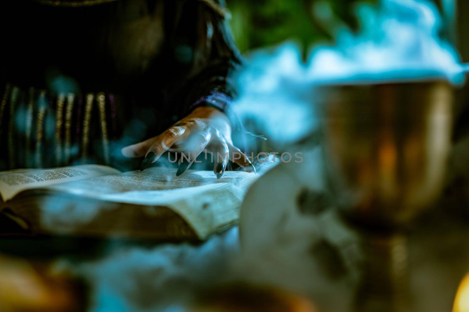 Close-up of a witch's hand with pointed black nails on smoky book of magic recipe and with a magic wand.