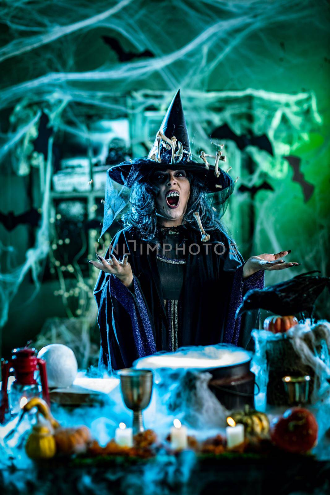 Witch with awfully face in creepy surroundings and smoky green background talks magic words to bones around her hat, above boiling cauldron. Halloween concept.