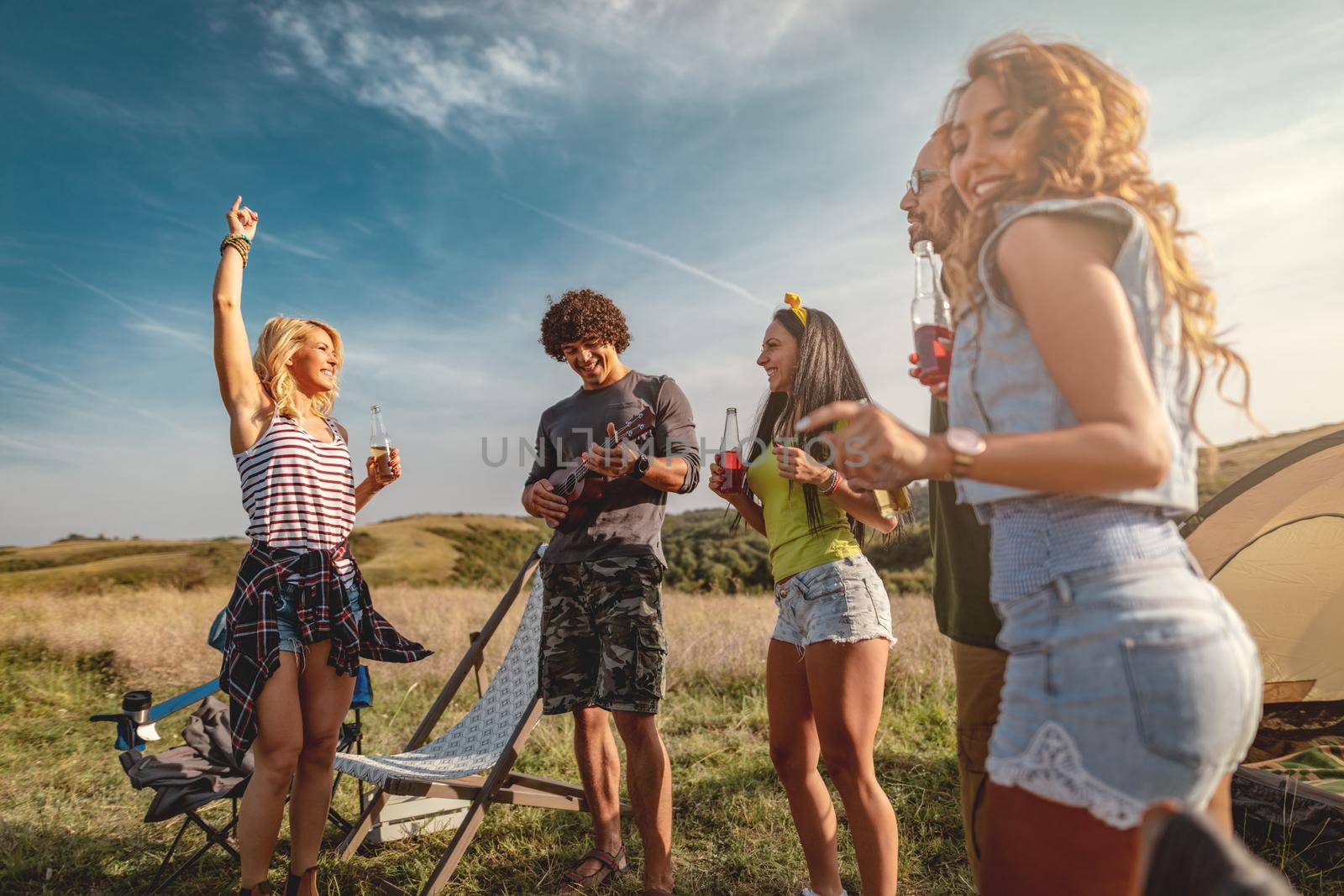 Young people have a good time in camp in nature. They're resting, laughing and singing with music from ukulele, happy to be together.