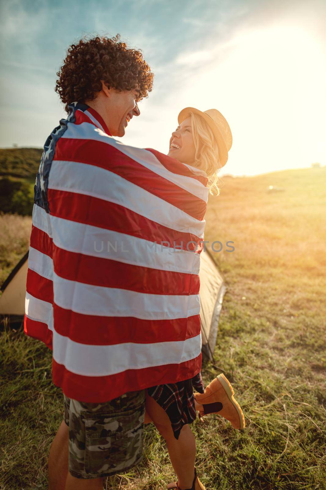 Happy young couple enjoys a sunny day in nature. They're hugging each other, wrapped in american flag in front a campsite tent. 