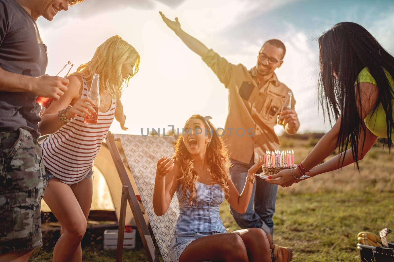 Young people have a good time in camp in nature. They're celebrating a birthday, laughing and greeting to their friend with birthday cake, happy to be together.