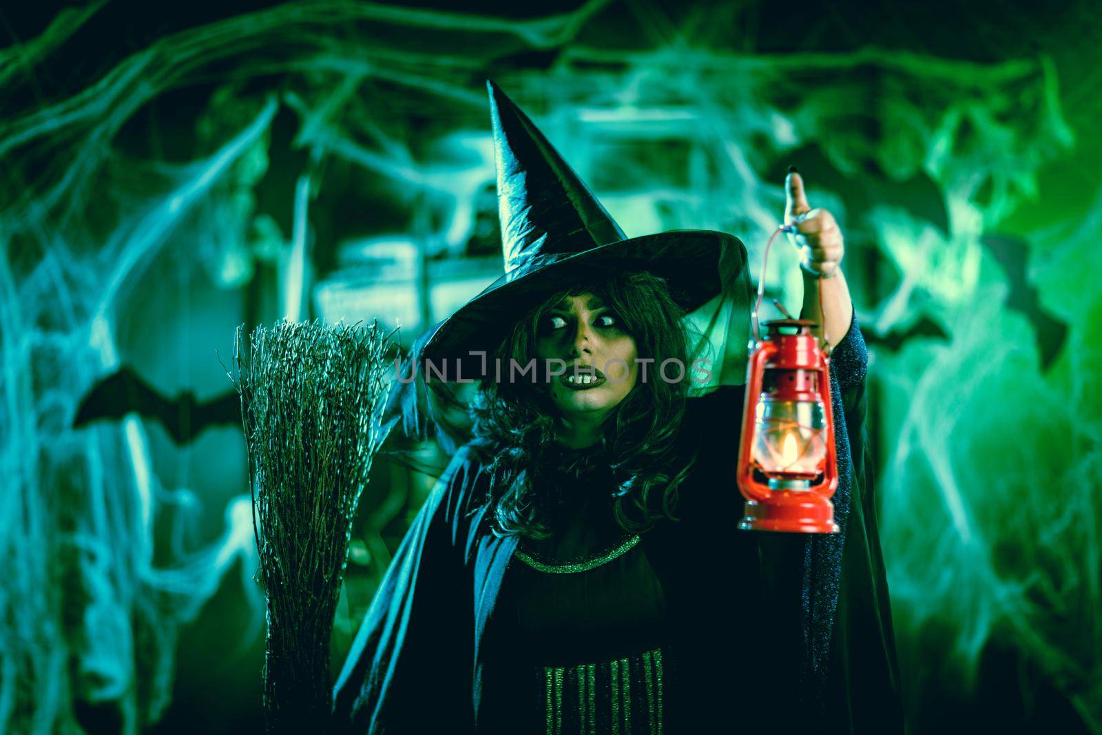 Portrait of witch with awfully face and lighted lantern in her hand in creepy foggy surroundings sends evil.