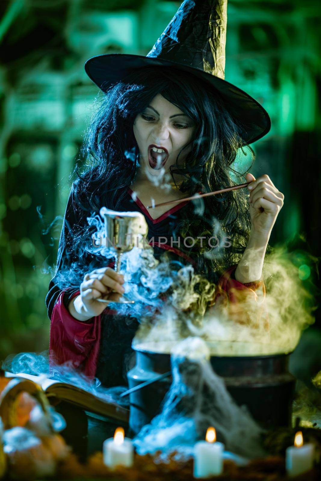 Young witch with seriously face in creepy surroundings and smoky green background holds a goblet with magic potion. 