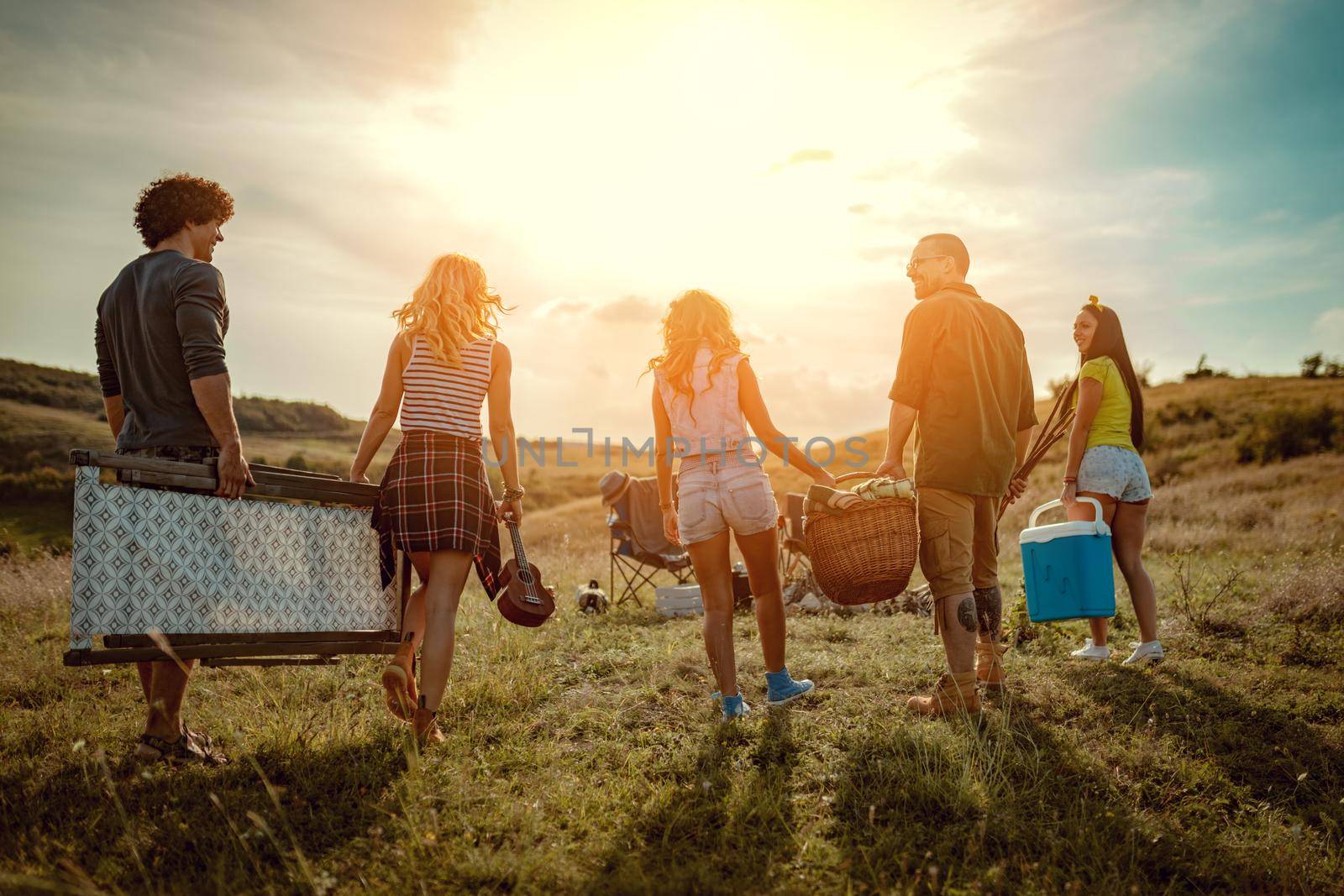 The young friends are preparing for camping. They're holding a resting chair, picnic basket, and other necessaries and searching for a suitable place in a sunset.