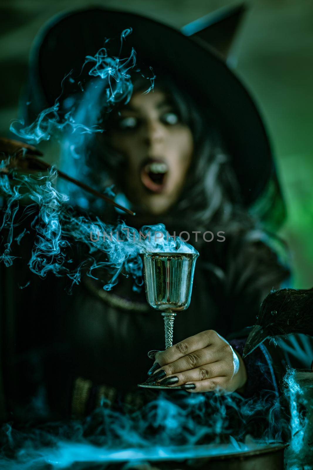 Portrait of witch with awfully face in creepy surroundings and smoky green background drinks magic potion from the goblet.