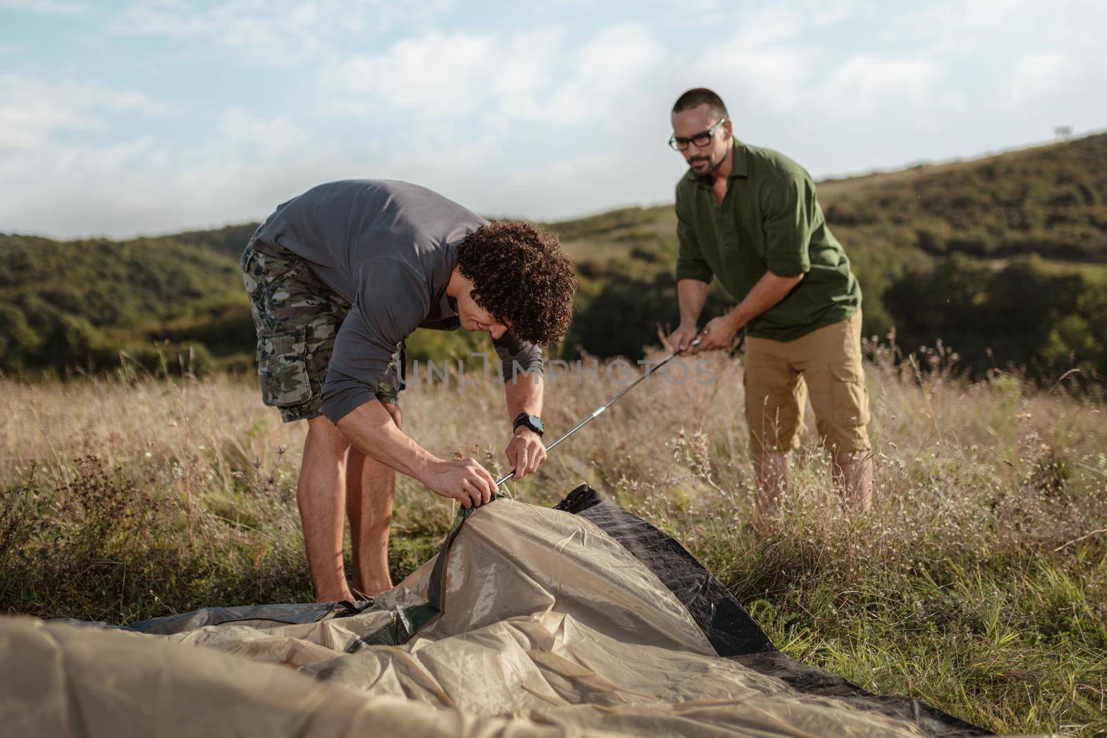 The young happy friends are preparing for camping. They're installing a tent on a suitable place in a meadow.