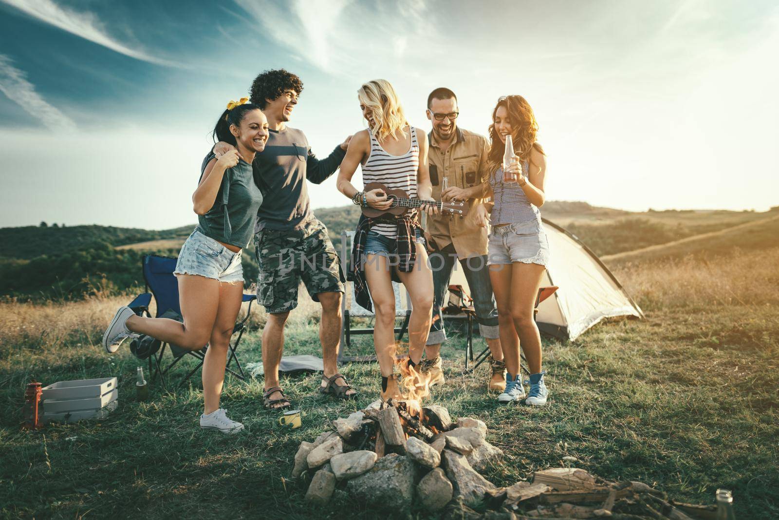 Happy young friends enjoy a sunny day at the mountain. They're laughing and singing with music from ukulele by the bonfire near tent.