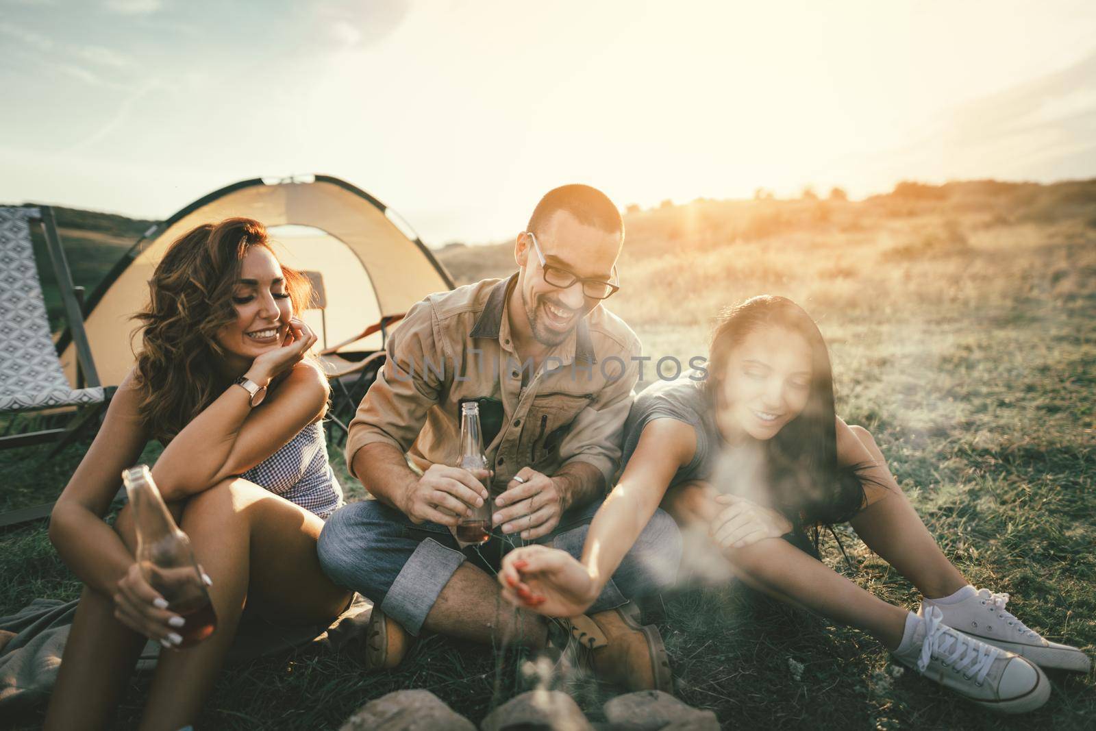 Happy young friends enjoy a sunset at the mountain. They're laughing and hanging out by the bonfire near tent.