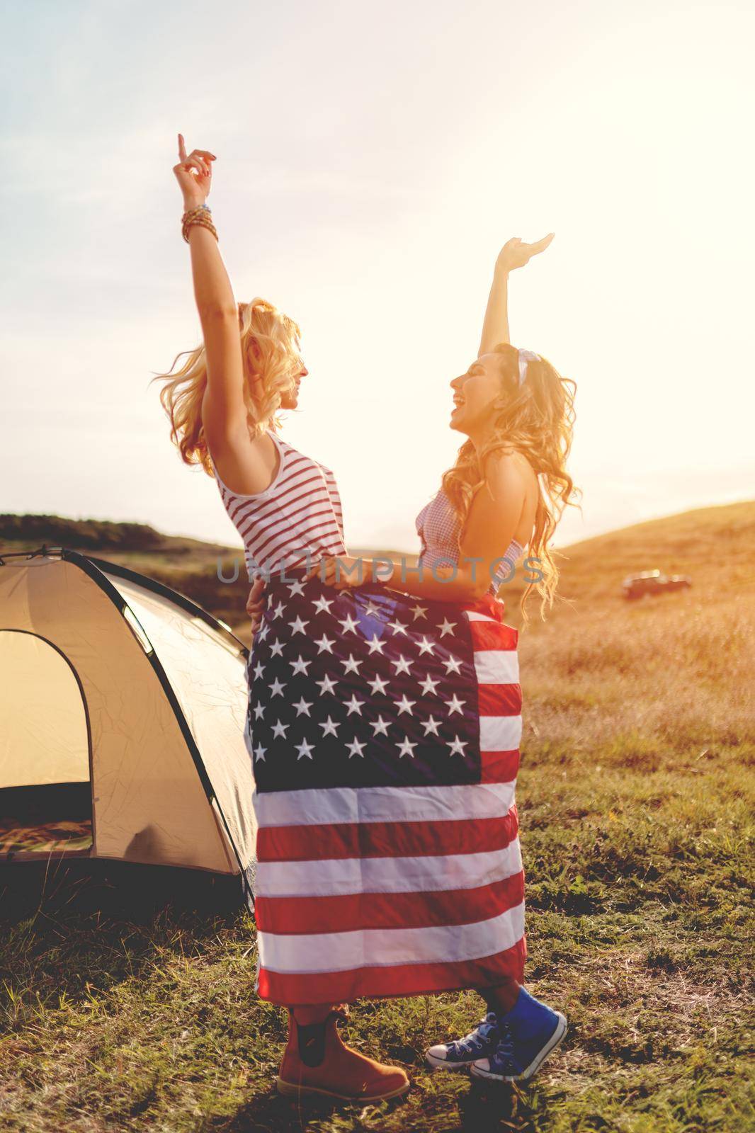 Happy young lesbian girl enjoys a sunny day in nature. They're wrapped in american flag in front a campsite tent and dancing. 