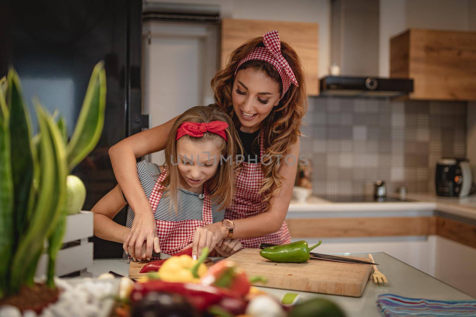 Cute little girl and her beautiful mom are cutting vegetables and smiling while cooking in kitchen at home.