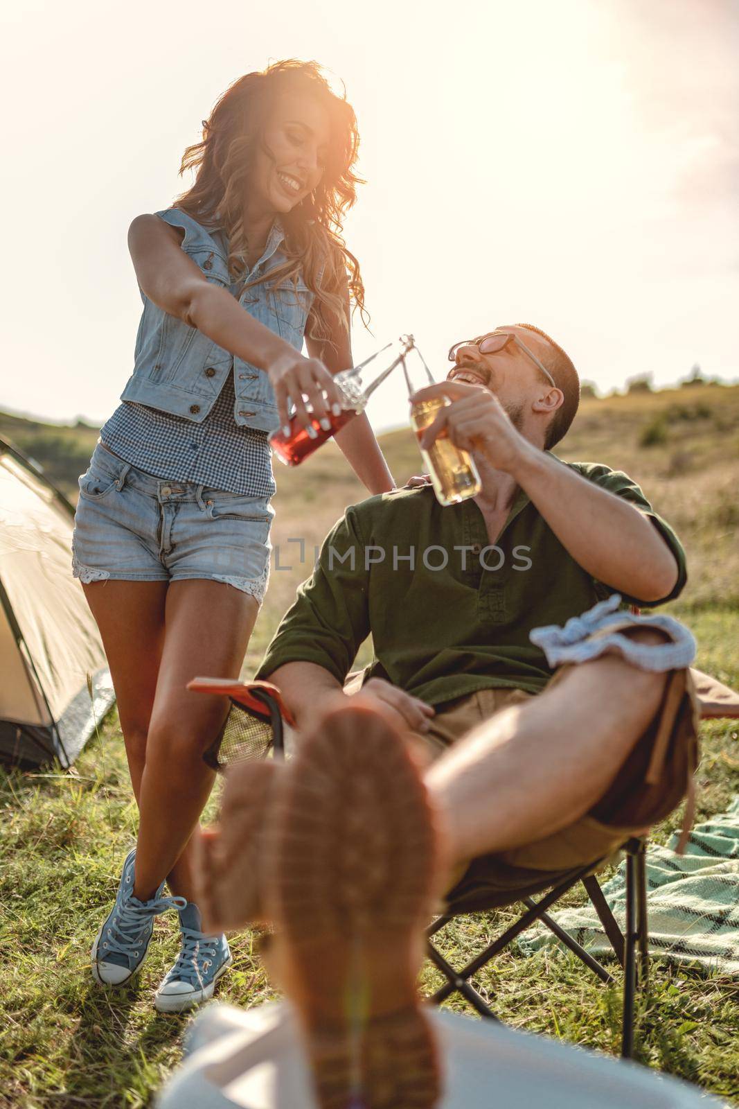 Happy young couple enjoys a sunny day in nature. They're clinking beer bottles in front a campsite tent. 