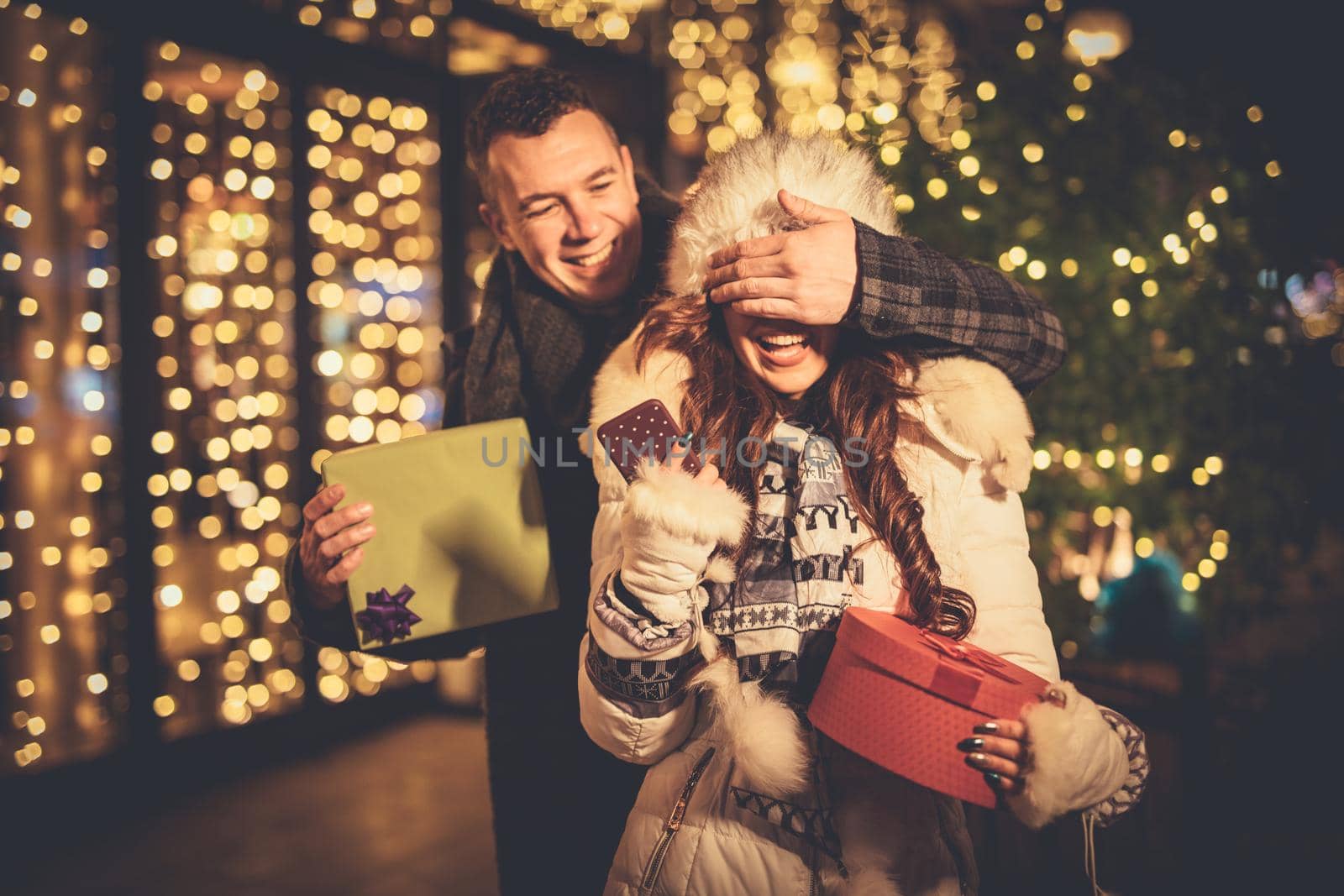 Young couple in the city centre with holiday's brights in background. Smiling man covering the eyes of happy surprised girlfriend in a Christmas night.