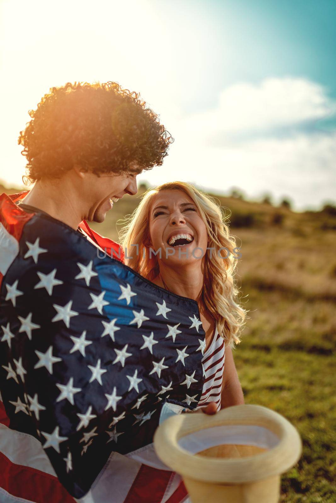 Happy young couple enjoys a sunny day in nature. They're hugging each other, wrapped in american flag. 