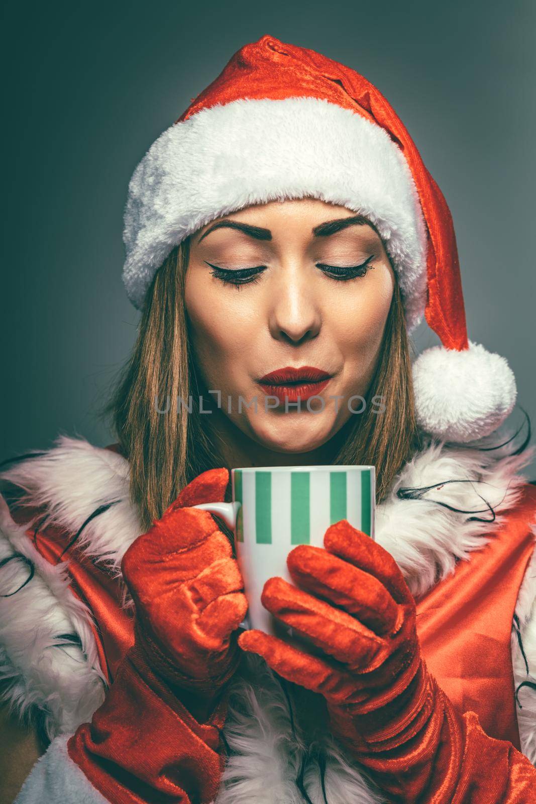 Portrait of a beautiful young smiling woman in Santa Claus costume. She is thinking and holding cup of hot tea.