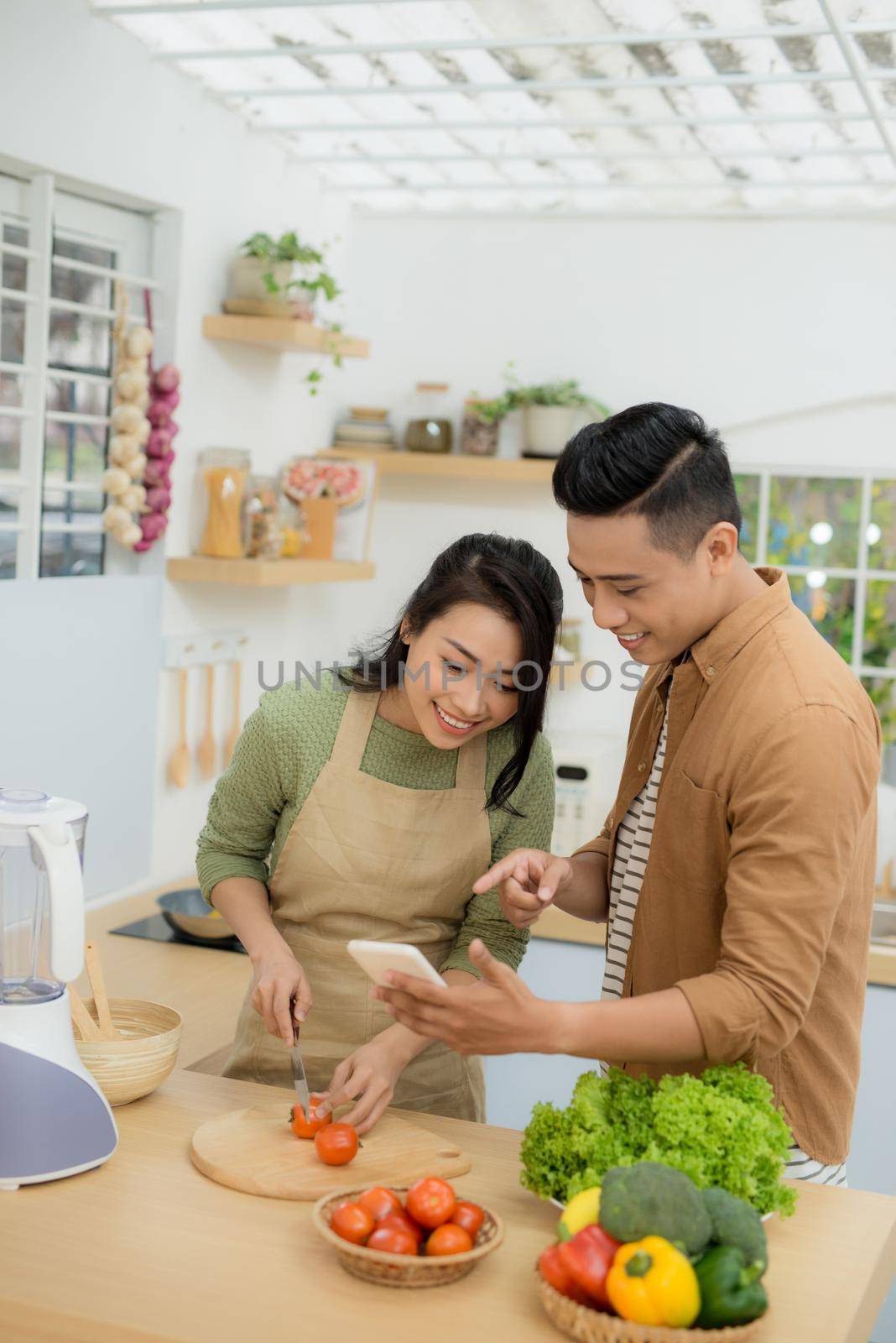 Beautiful young couple is using a cellphone and smiling while cooking in kitchen at home by makidotvn