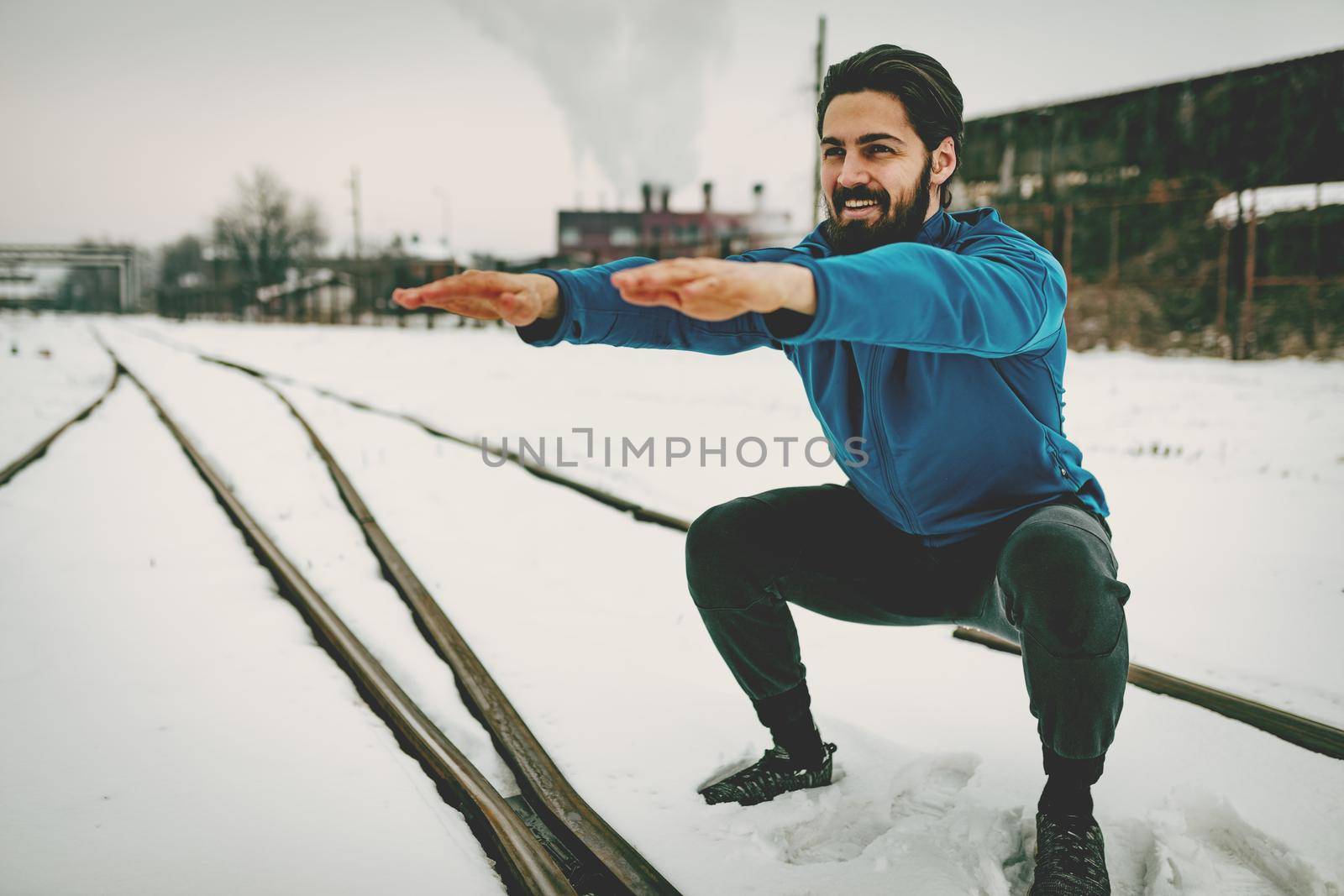 Active young man crouching and doing exercises in the public place among old railroad during the winter training outside in. Copy space.